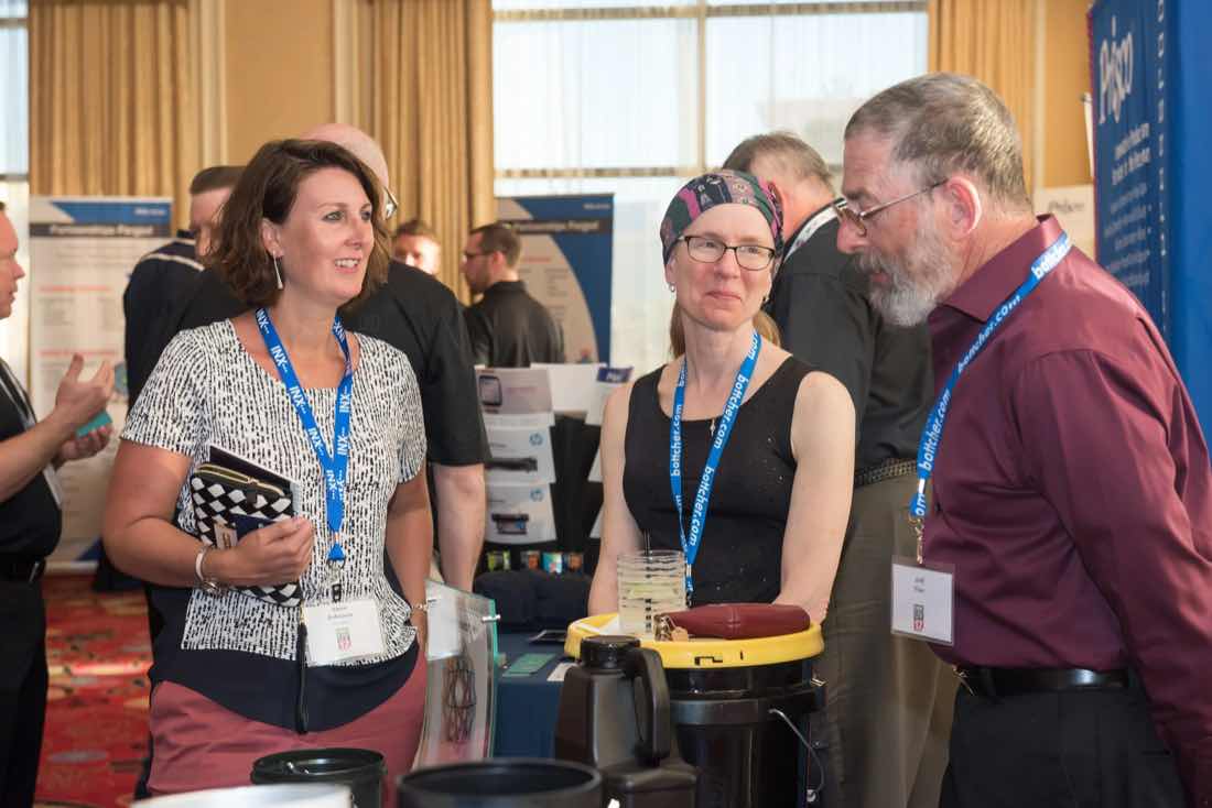 Three attendees with badges chatting beside sponsor displays at the conference