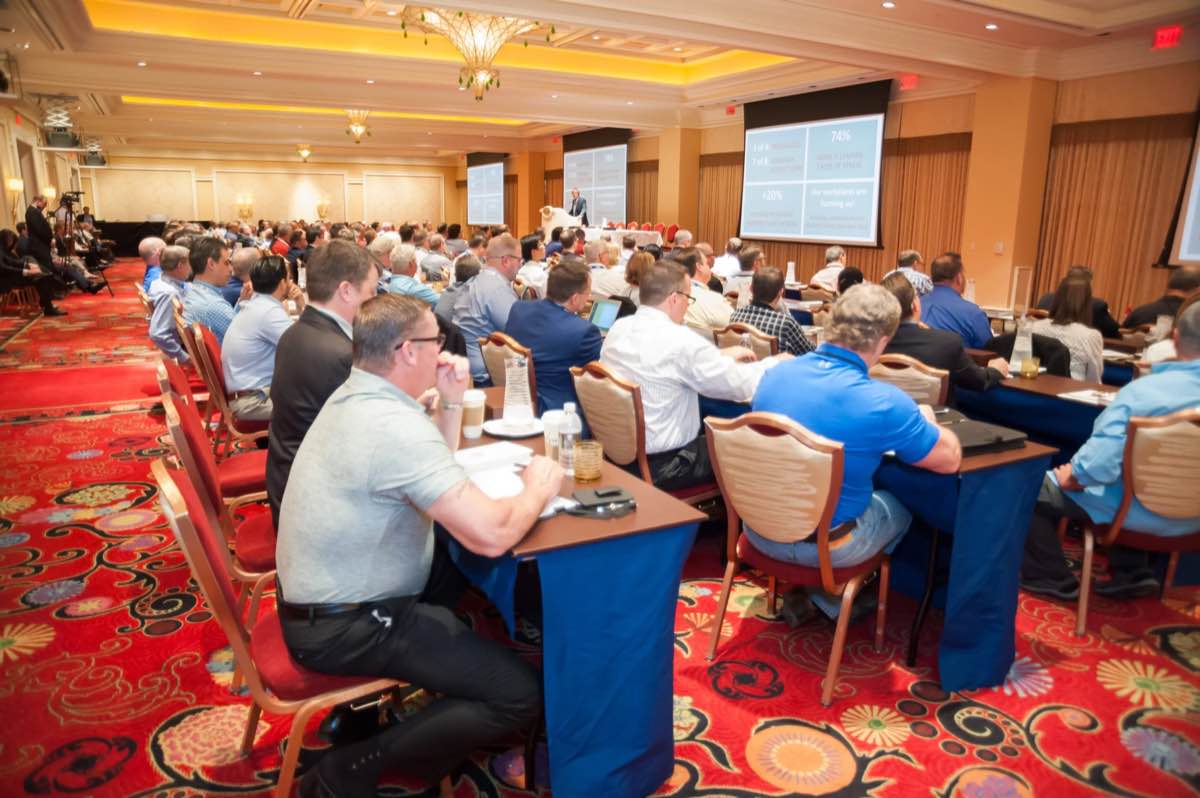 Full ballroom of conference attendees at long tables watching a presentation on two screens
