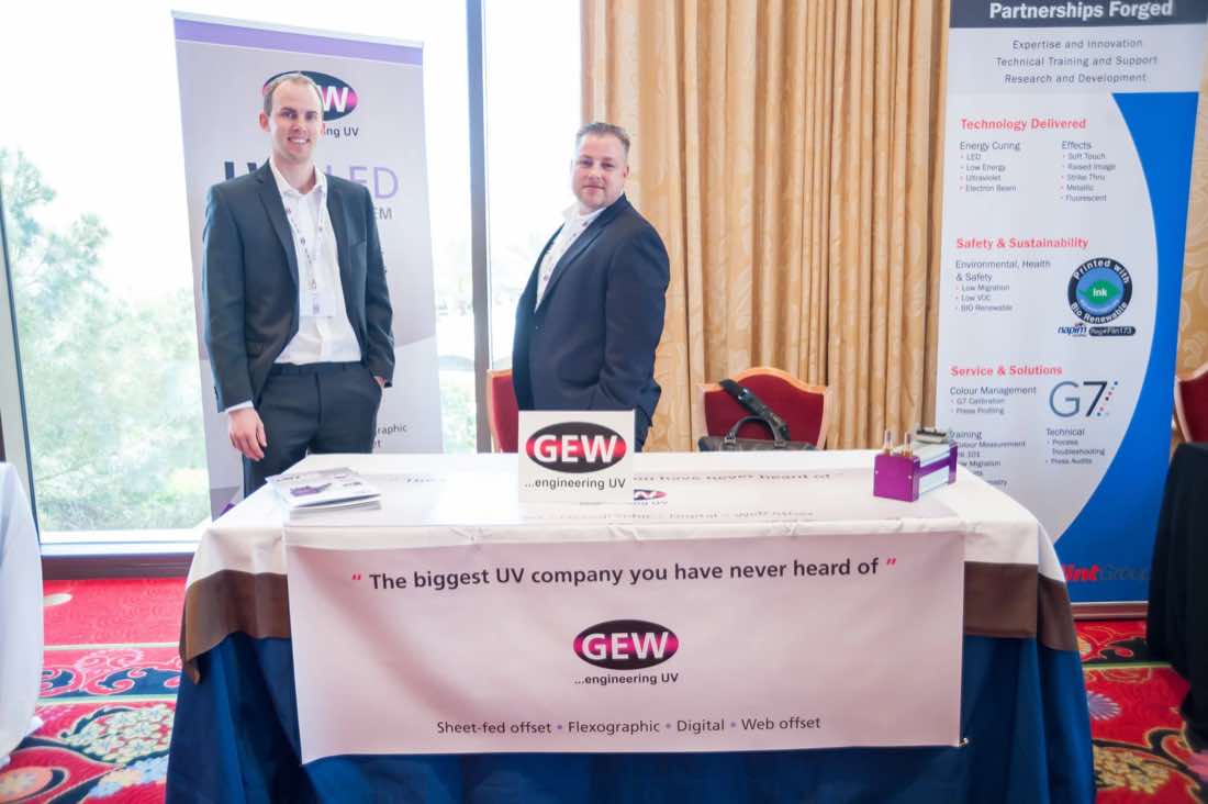 Two exhibitors standing at a GEW UV sponsor table with banner and literature