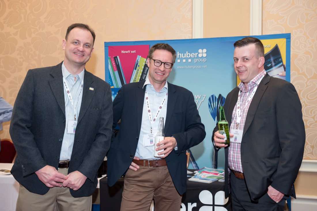 Three men with drinks in front of a hubergroup ink sponsor display