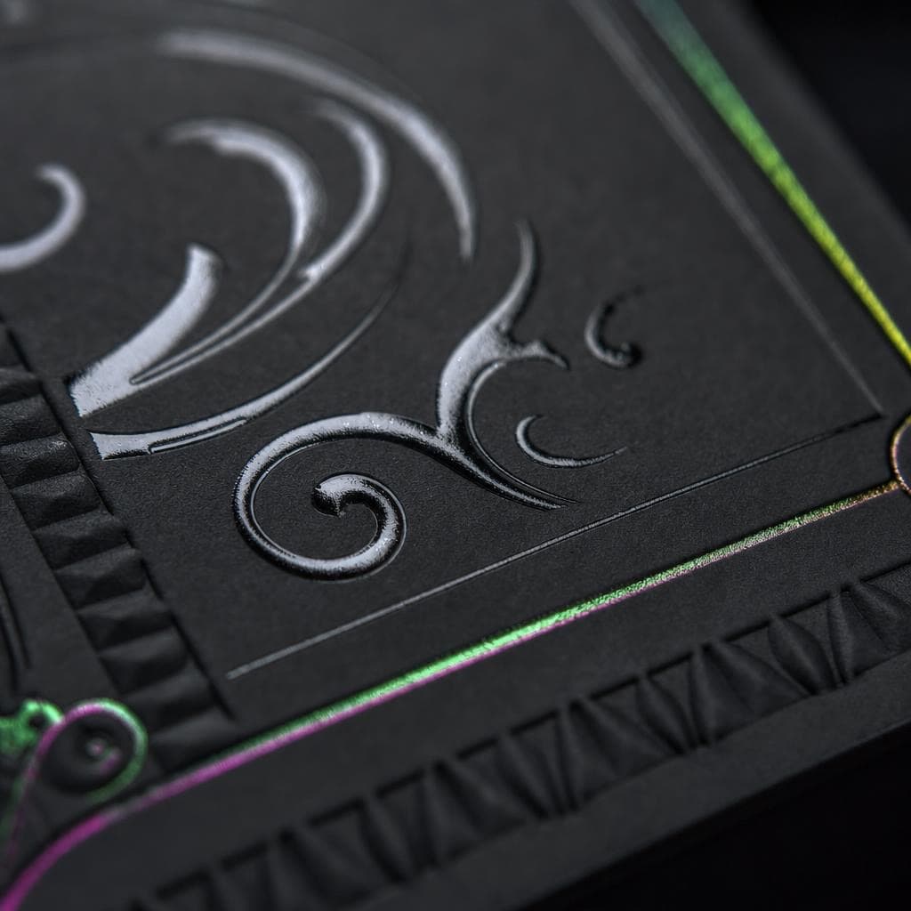 Macro of a blind-embossed flourish and a green-to-magenta spectrum foil keyline with spot gloss over soft-touch matte black stock