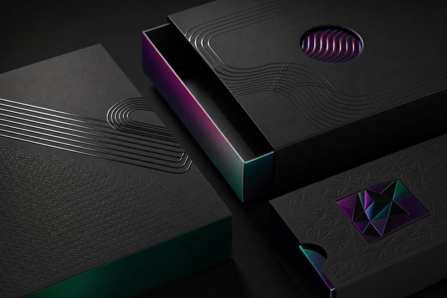 Matte black rigid boxes finished with debossed linework, iridescent green-to-magenta foil edges and a faceted foil emblem, lit low-key in a dark studio