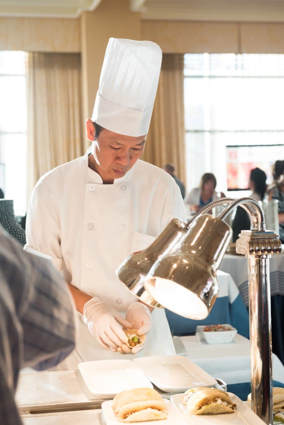 Chef in whites and toque hand-finishing plates under a heat lamp at a buffet station