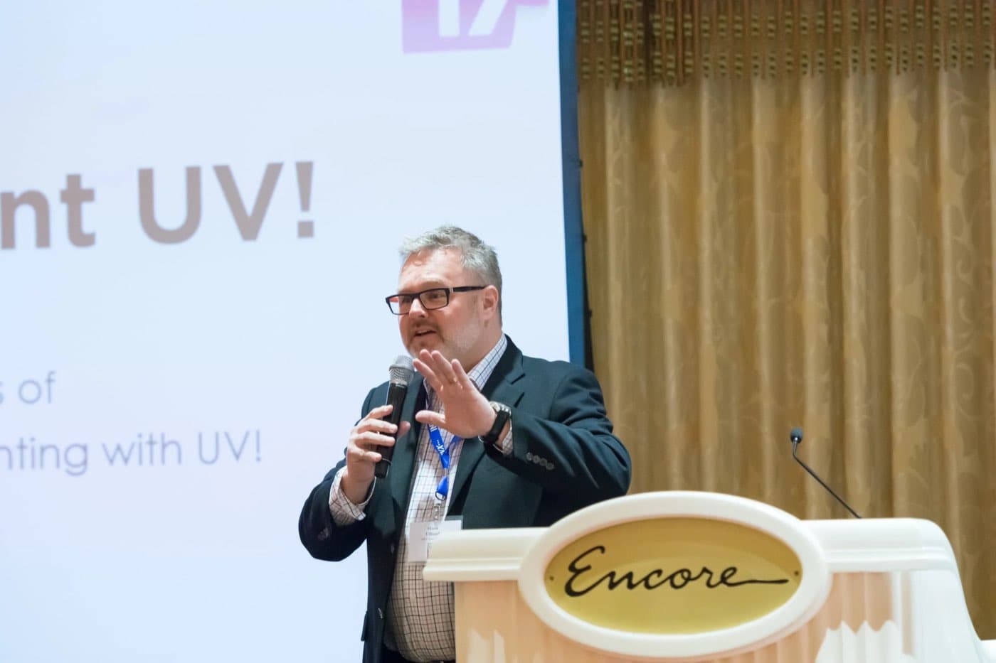 Speaker presenting at a podium beside a projected slide about printing with UV